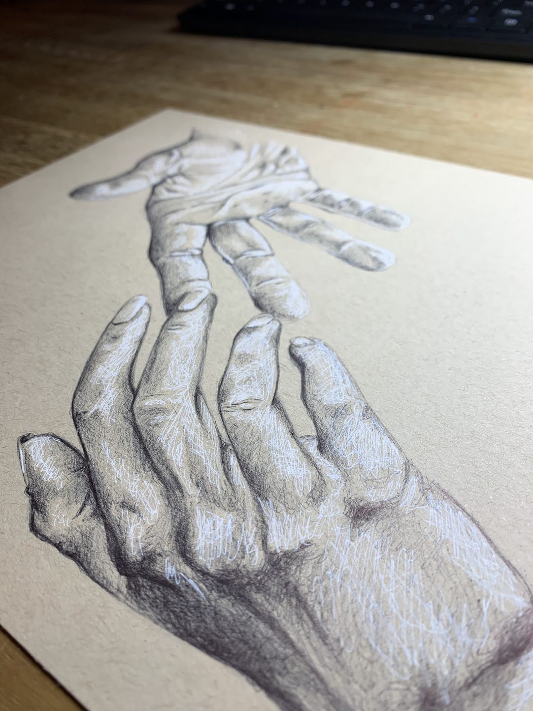 A drawing in pen and white on toned paper of two hands reaching toward each other, photographed at an angle on a desk